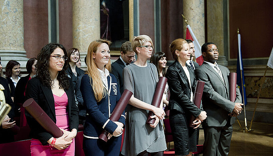 5 Absolvent*innen im Festsaal der Universität mit ihren frisch erhaltenen Diplomen.